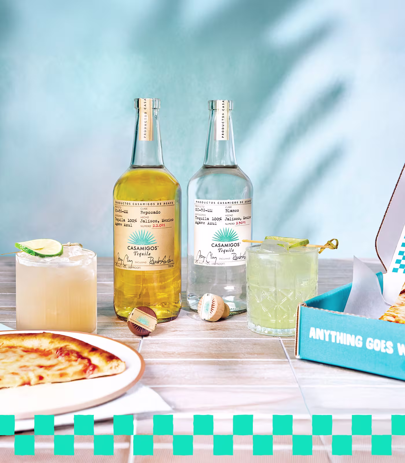 Two bottles of Casamigos tequila, two cocktails with lime, and a pizza in a box on a light wooden table with a blue background.