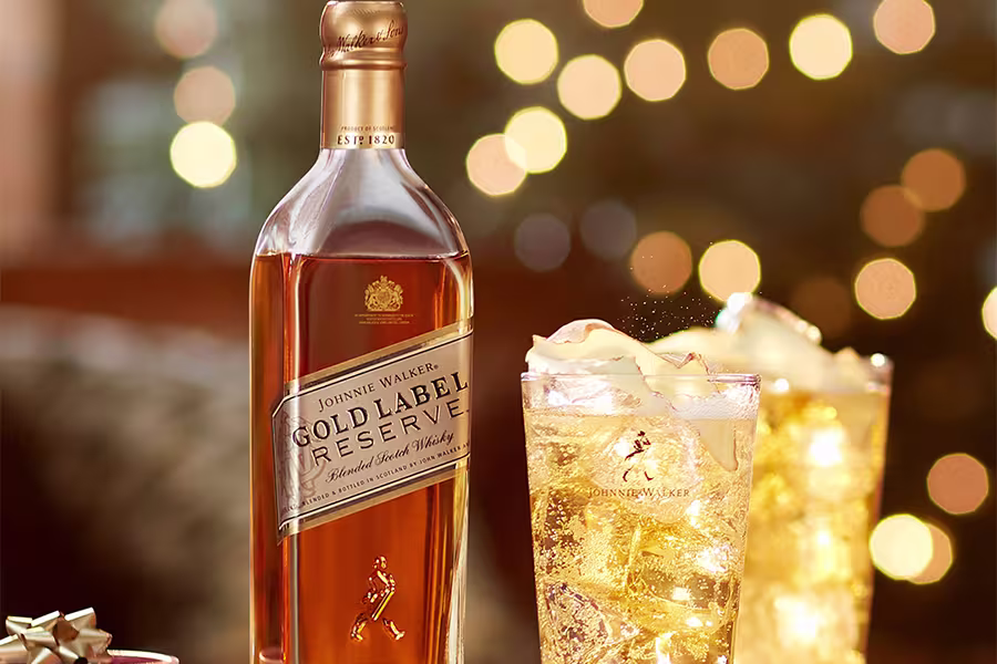 A bottle of Johnnie Walker Gold Label Reserve whisky stands next to two glasses filled with a golden drink and ice, with blurred golden lights in the background.