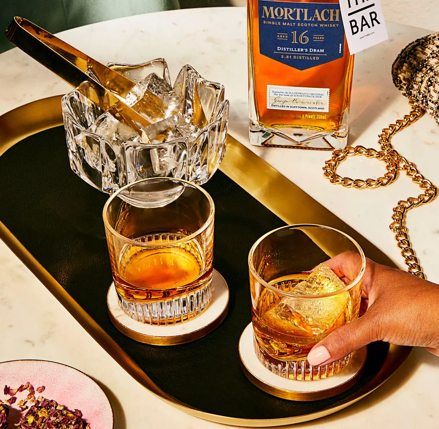 A hand holds a glass of whisky with ice on a tray, next to another glass, an ice bucket with tongs, and a bottle of Mortlach 16 Year Old Single Malt Scotch Whisky.