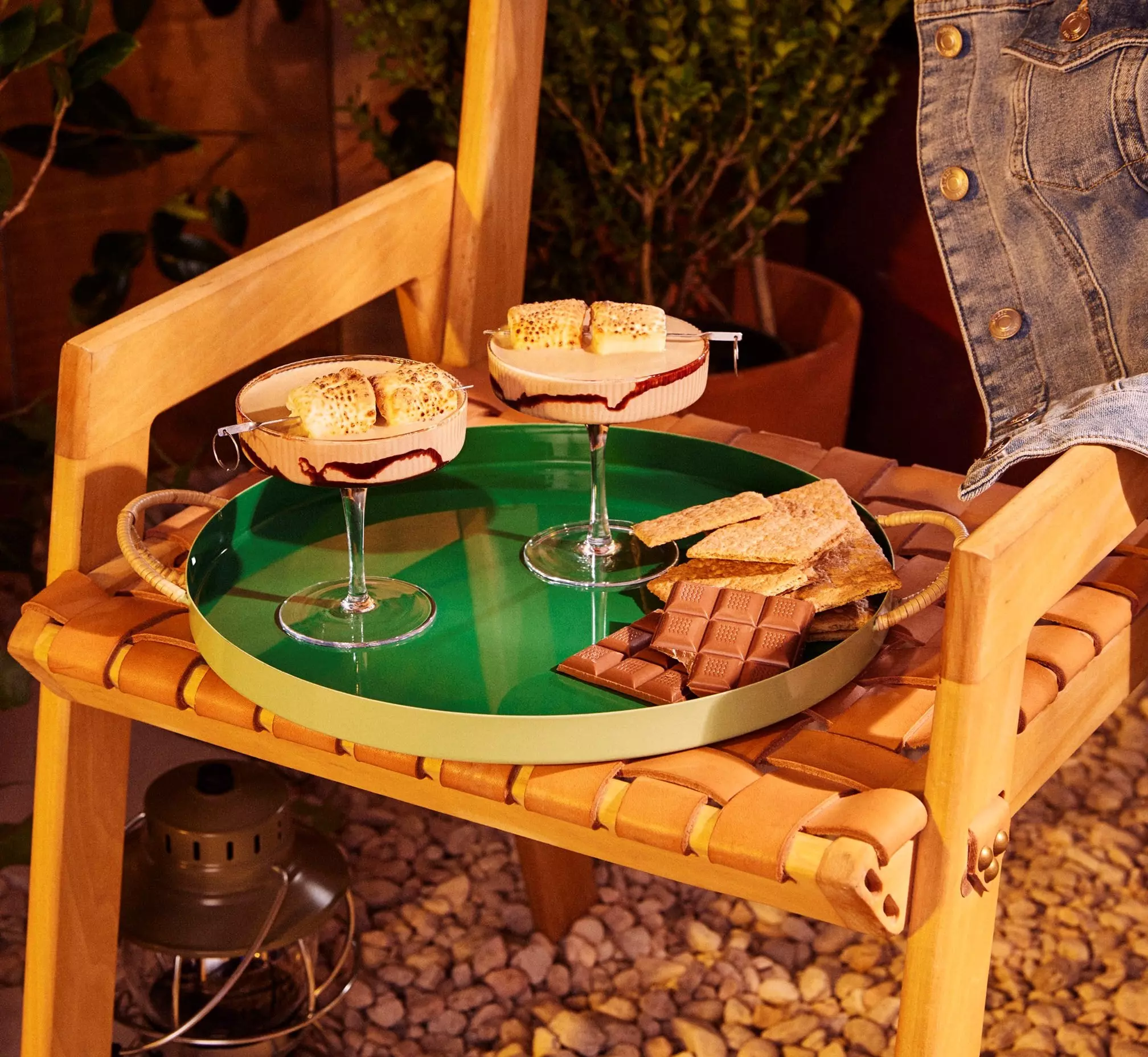 A green tray on a wooden chair holds two glasses of dessert topped with toasted marshmallows, chocolate bars, and graham crackers.