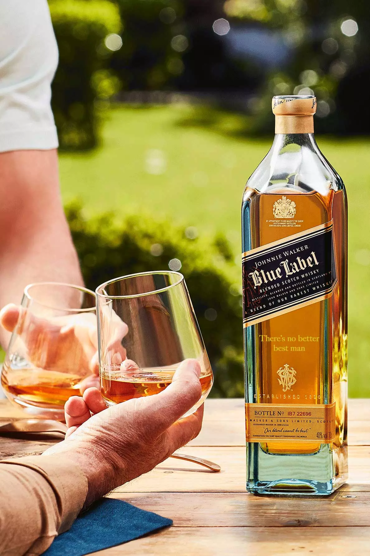 A bottle of Johnnie Walker Blue Label whisky sits on a wooden table outdoors, with two people holding glasses of whisky in the background.