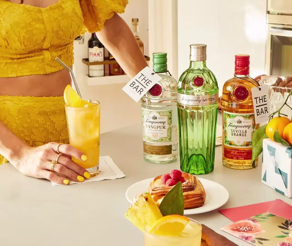 A person in a yellow outfit holds a cocktail beside three bottles of Tanqueray gin on a counter with pastries, fruit, and bar tags.