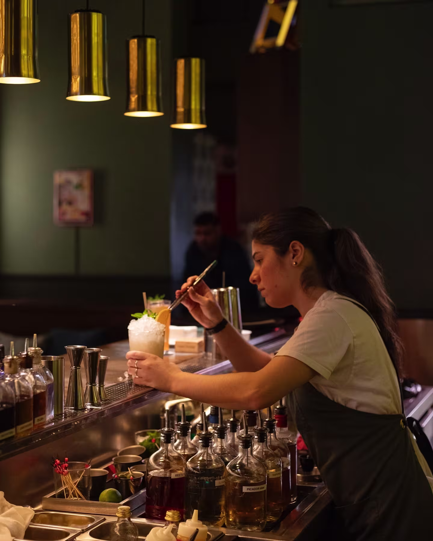 Bartender en El Chiringuito de Casa Bonay preparando cóctel con hielo picado y menta, rodeada de botellas y utensilios en barra iluminada.