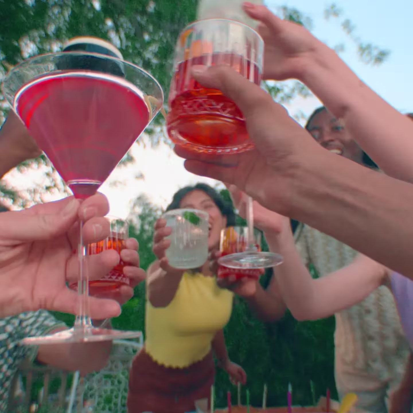 A group of people raise various drinks in a toast at an outdoor gathering, with greenery in the background.