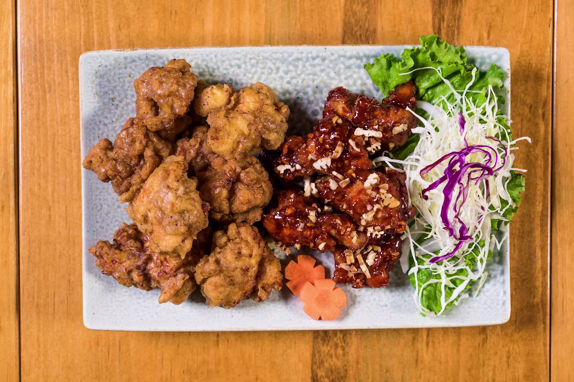 A rectangular plate with fried chicken pieces, glazed chicken pieces, shredded cabbage on lettuce, and two carrot slices on a wooden table.