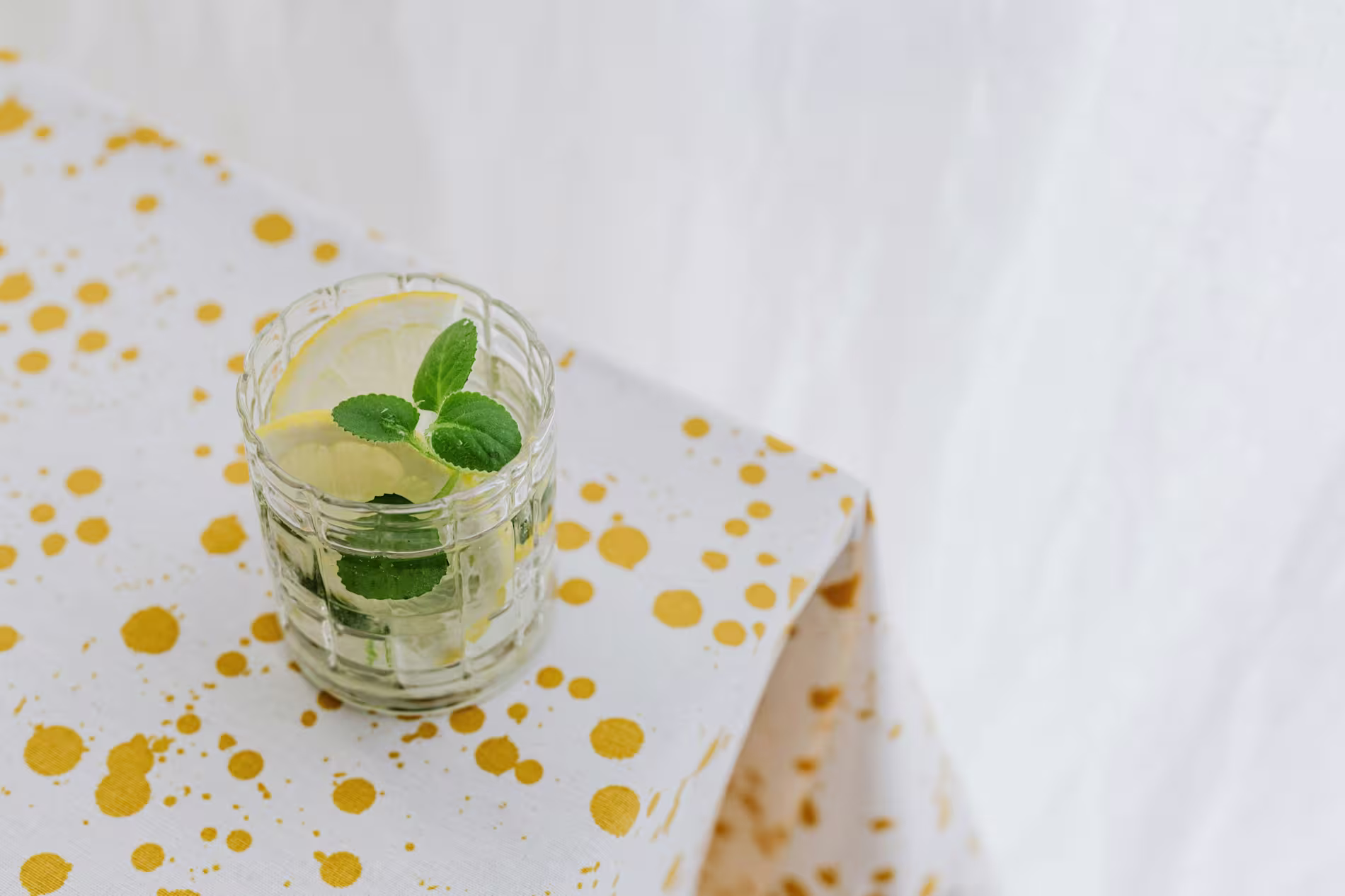 A glass of water with lemon slices and mint leaves sits on a white tablecloth with yellow splatter patterns.
