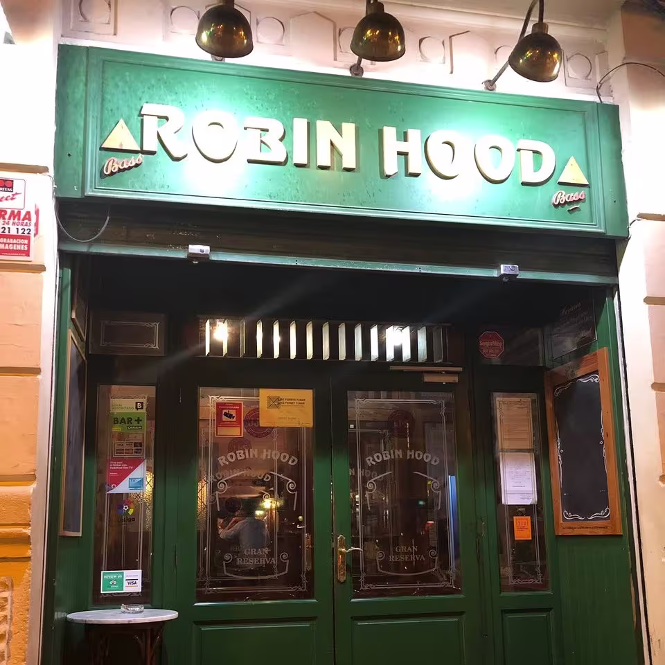 Entrada verde del pub Robin Hood con letras doradas, tres lámparas vintage y una mesa alta con cenicero en la puerta.