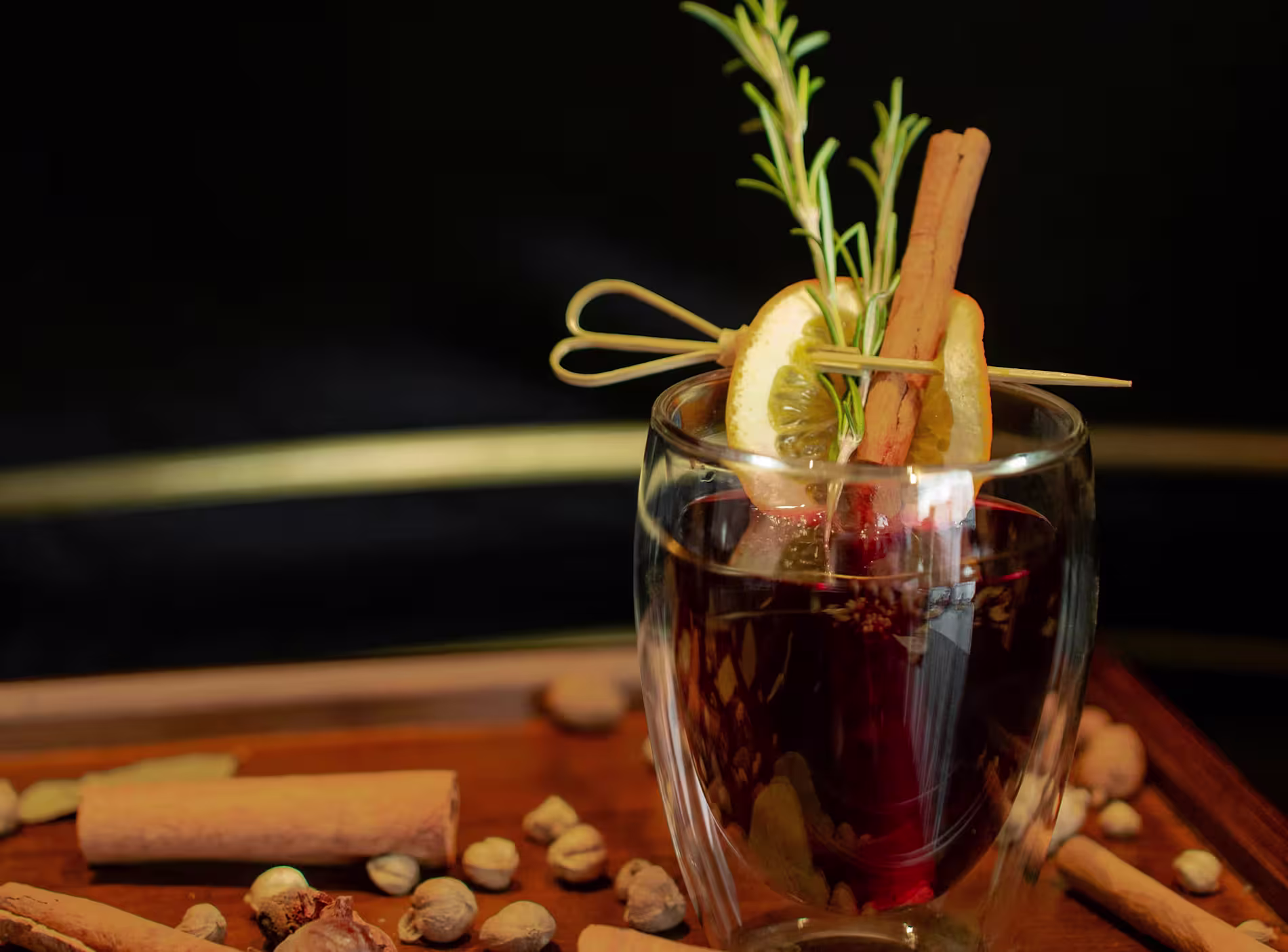 A glass of mulled wine garnished with a lemon slice, rosemary sprig, and cinnamon sticks, surrounded by whole spices on a wooden tray.