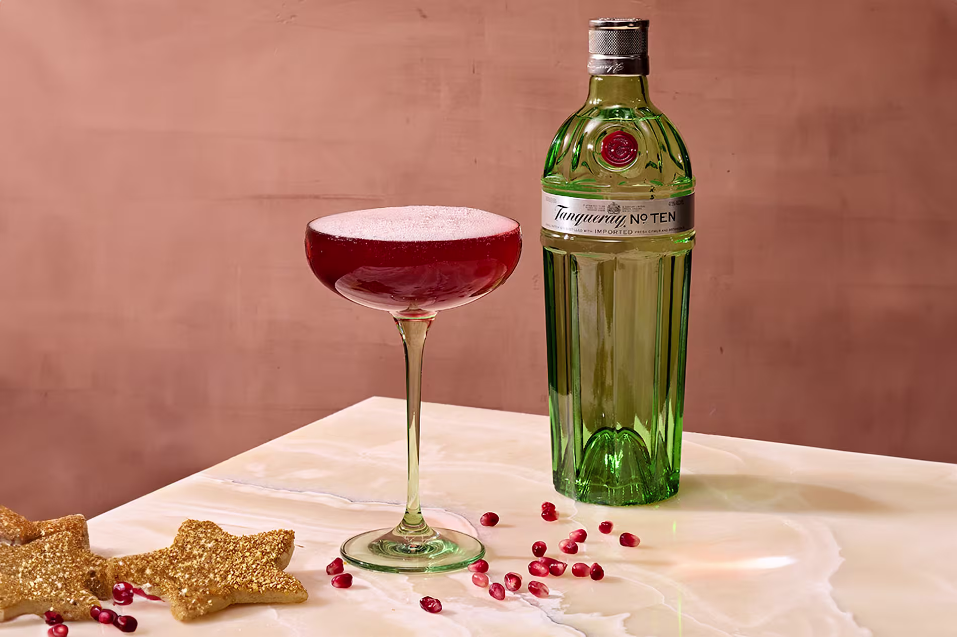 A green Tanqueray No. Ten gin bottle stands next to a coupe glass filled with a red cocktail and foam, on a marble surface with pomegranate seeds and glittery gold star decorations.