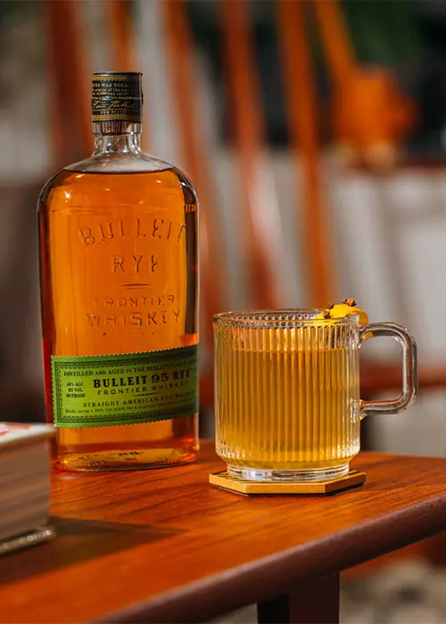 A bottle of Bulleit Rye Frontier Whiskey next to a clear glass mug filled with a light amber drink, garnished with an orange peel, on a wooden table.