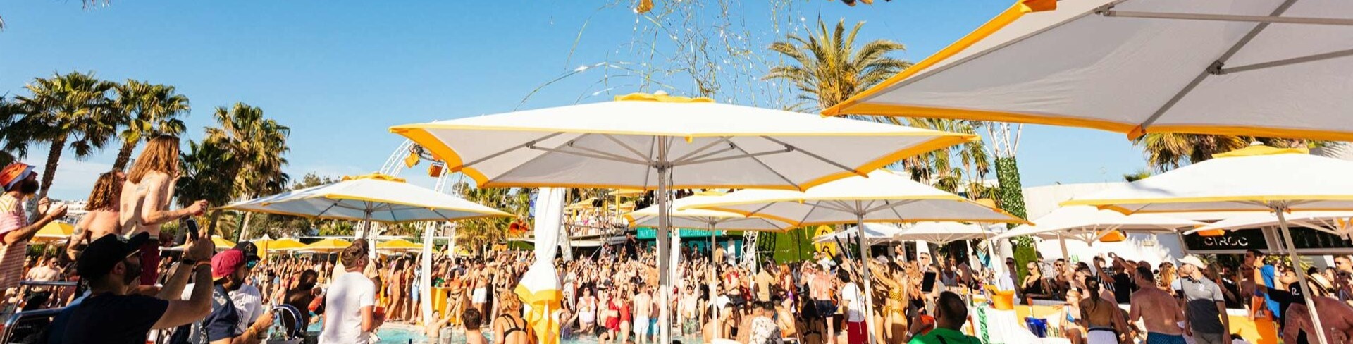 Fiesta en O Beach Ibiza con multitudes disfrutando junto a la piscina, sombrillas amarillas y un espectáculo aéreo con decoraciones vibrantes.