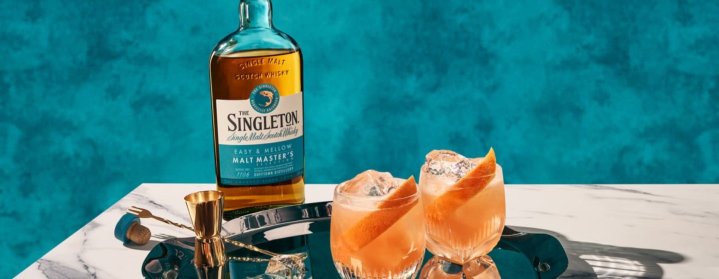 O sticlă de The Singleton Malt Master's se află pe o masă de marmură, alături de două cocktailuri pe o tavă de metal