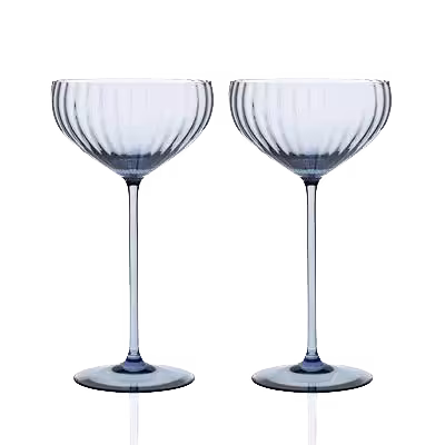 Two clear, ribbed glass coupe cocktail glasses with long stems are standing side by side against a plain background.