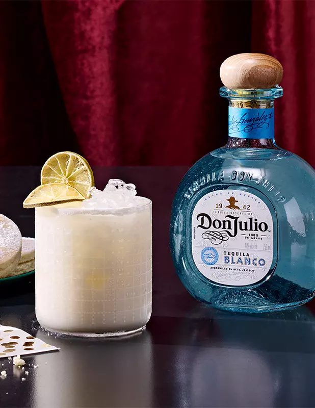 A glass of iced cocktail garnished with lime sits next to a bottle of Don Julio Tequila Blanco on a dark table with a red curtain backdrop.