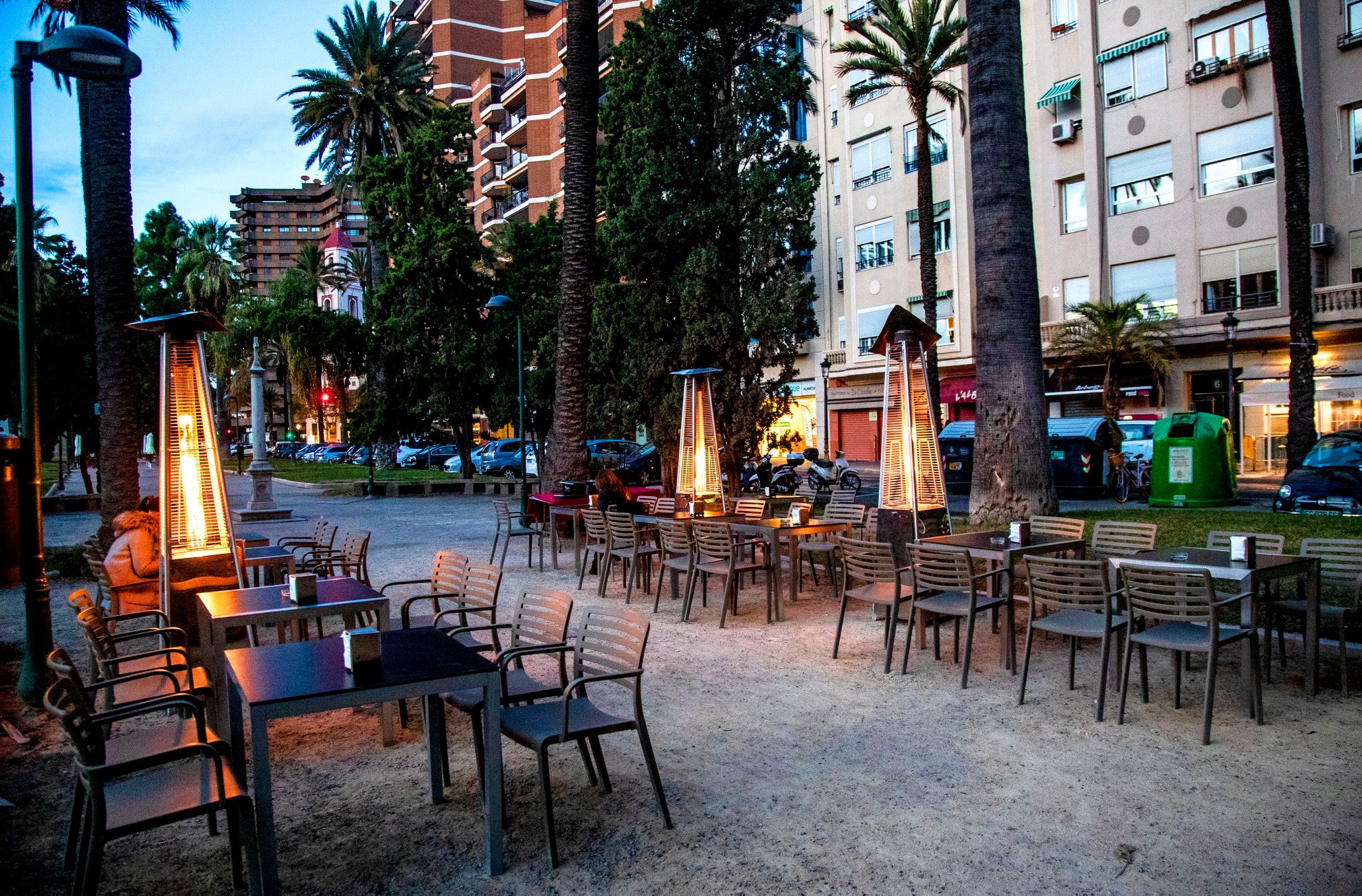 Terraza de Café Alameda al anochecer, con estufas exteriores encendidas entre palmeras, mesas distribuidas en la plaza y ambiente relajado junto a la ciudad.