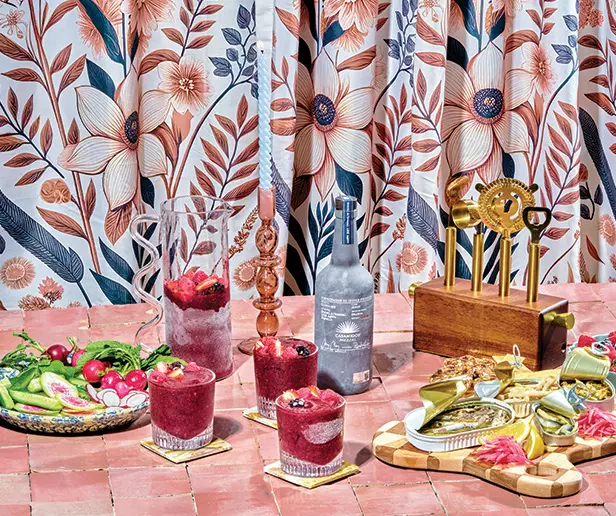 A table with three berry drinks, a bottle of alcohol, a plate of vegetables, mixed bar tools, and a pitcher, set against a floral curtain background.
