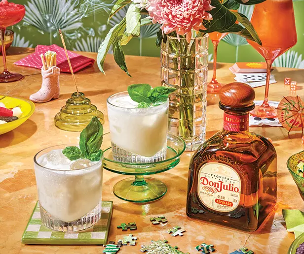 A table set with two creamy cocktails, a bottle of Don Julio tequila, a flower vase, a red drink, snacks, and puzzle pieces.
