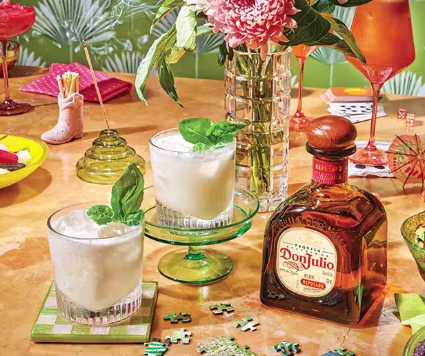 A table set with two creamy cocktails, a bottle of Don Julio tequila, a flower vase, a red drink, snacks, and puzzle pieces.