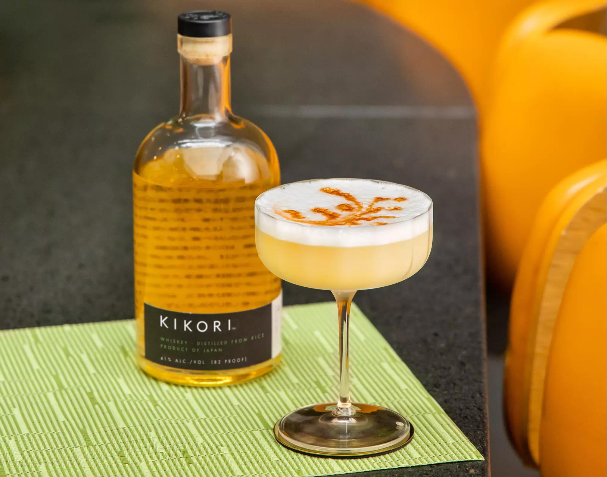 A bottle of Kikori whiskey and a coupe glass with a frothy cocktail topped with an orange garnish sit on a green mat on a dark counter.
