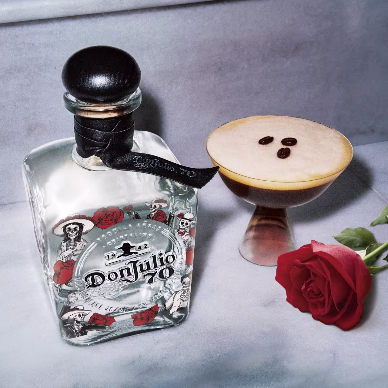 A bottle of Don Julio 70 tequila with Day of the Dead-themed label, a cocktail garnished with three coffee beans, and a red rose on a marble surface.