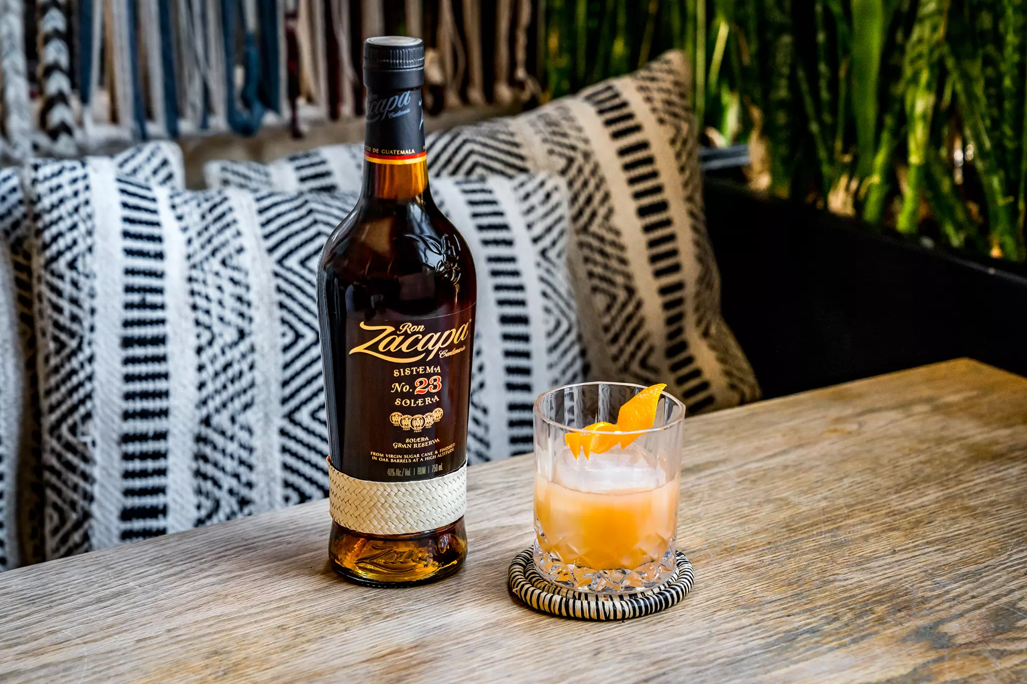 A bottle of Ron Zacapa rum stands next to a glass of cocktail with ice and an orange peel garnish on a wooden table, with cushions and greenery in the background.