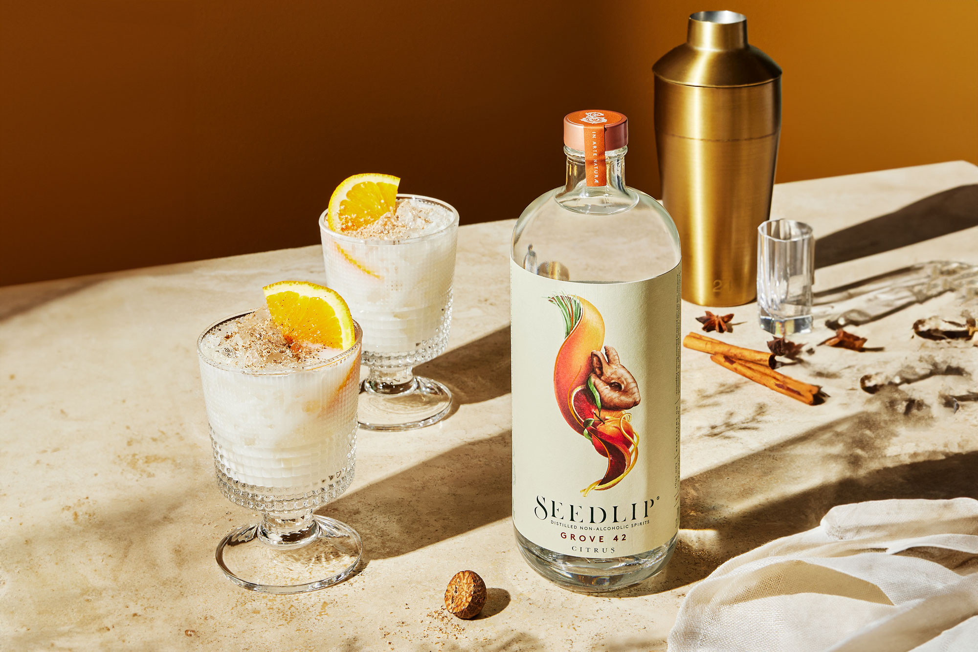 A bottle of Seedlip Grove 42, two cocktail glasses with creamy drinks garnished with orange slices, a cocktail shaker, and various spices on a beige surface.