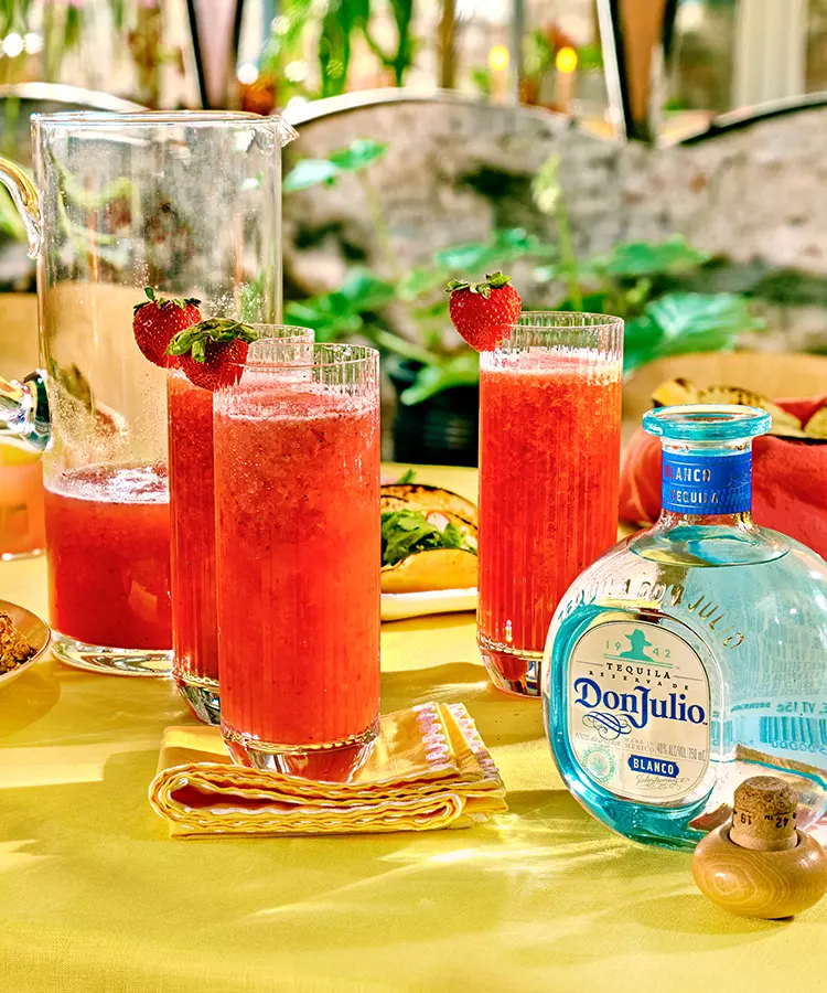 Three glasses of red strawberry cocktails with whole strawberries on the rim, a pitcher, and a bottle of Don Julio Blanco tequila on a yellow tablecloth.