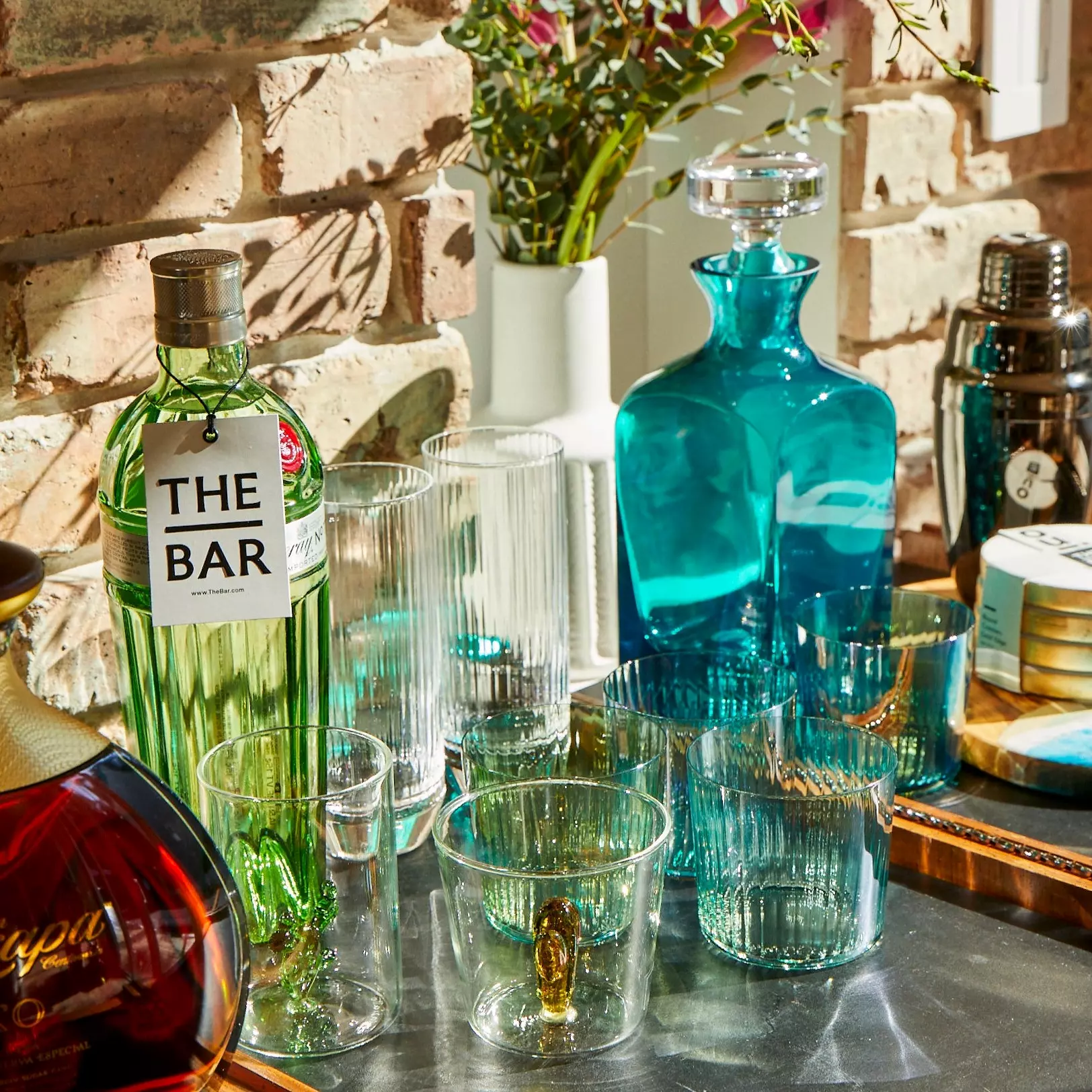 A collection of glassware, a green bottle labeled The Bar, a blue decanter, and cocktail accessories arranged on a table against a brick wall.