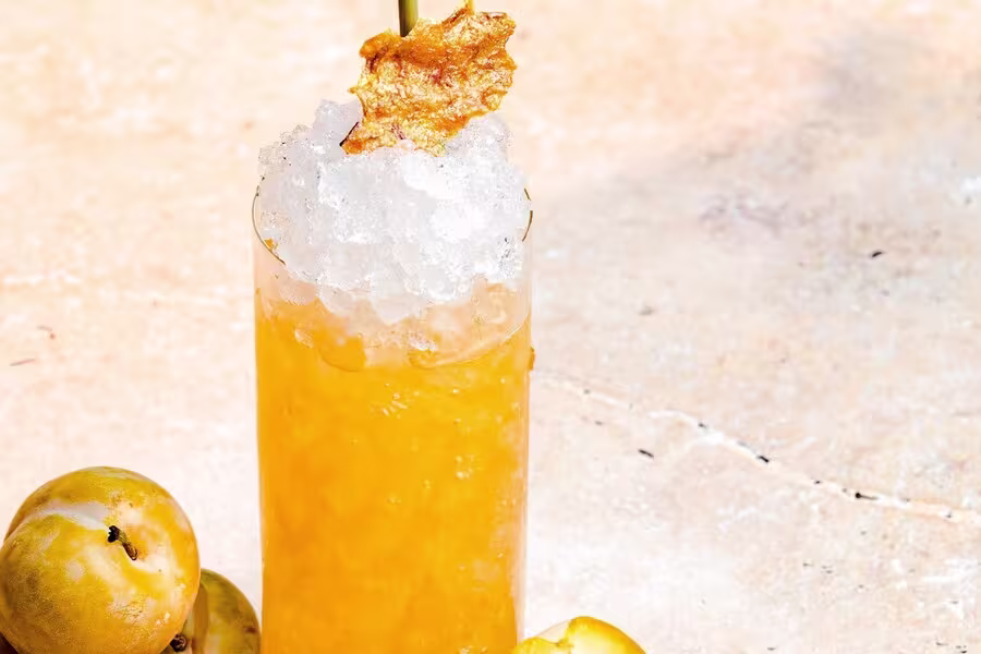 A tall glass of orange-yellow drink with crushed ice, garnished with a golden crisp, next to several yellow fruits on a light-colored surface.