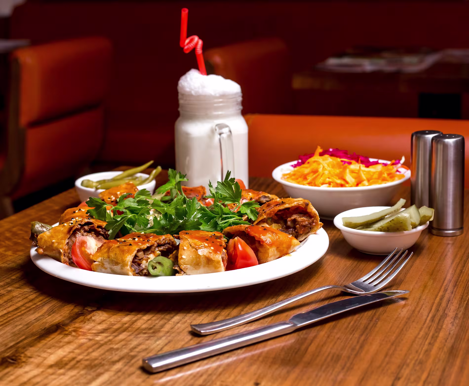 A plate of stuffed pastry rolls with greens and tomatoes, a glass of a white frothy drink, a side of salad, pickles, and salt and pepper shakers on a wooden table.