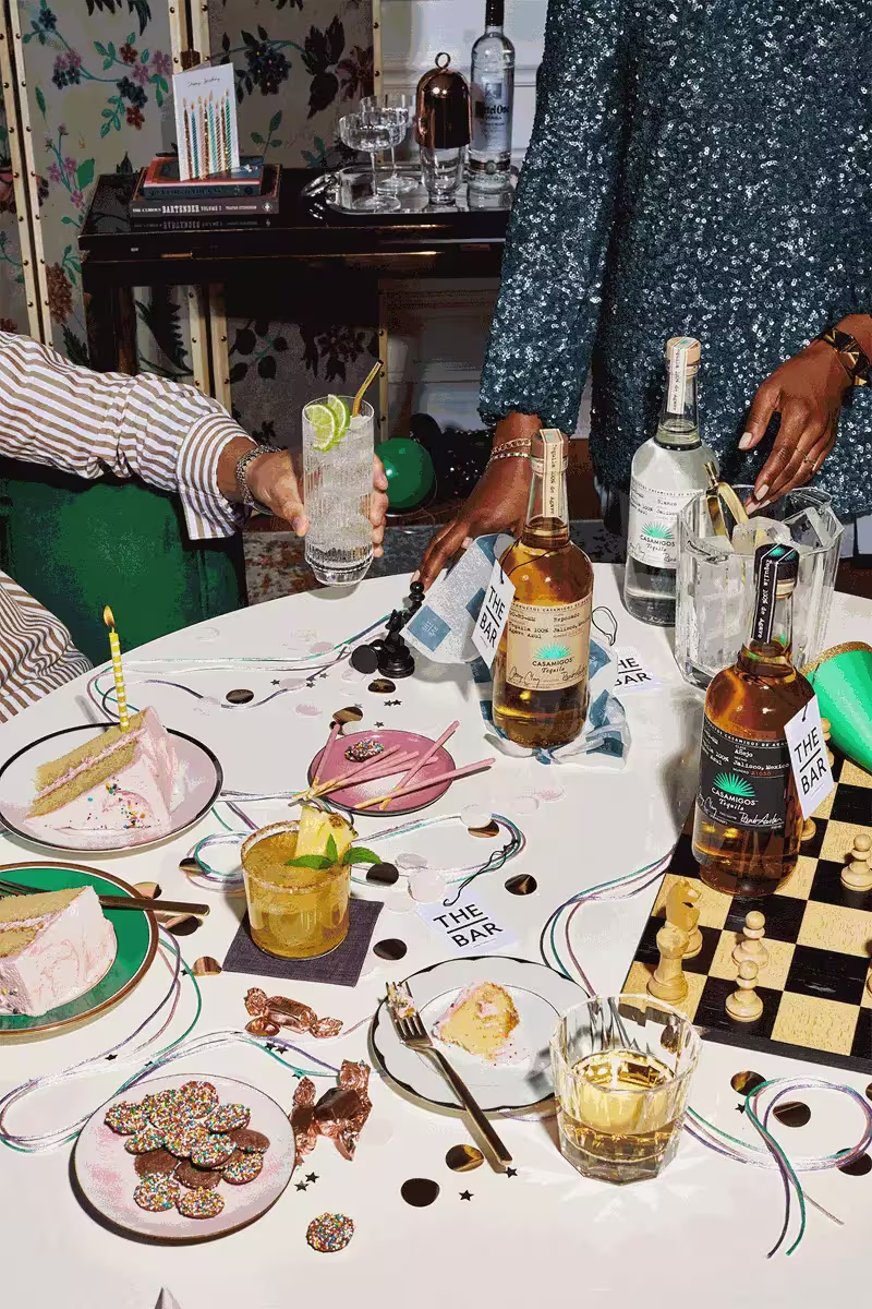 Two people at a decorated table with drinks, cake, party favors, and a chessboard. Bottles of spirits and glasses are arranged amid festive confetti and snacks.