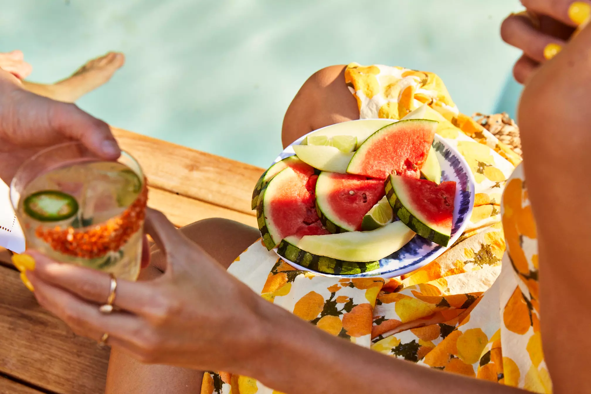 A person in a yellow floral dress holds a plate of watermelon and melon slices by a pool, while another hand offers a glass with a drink.