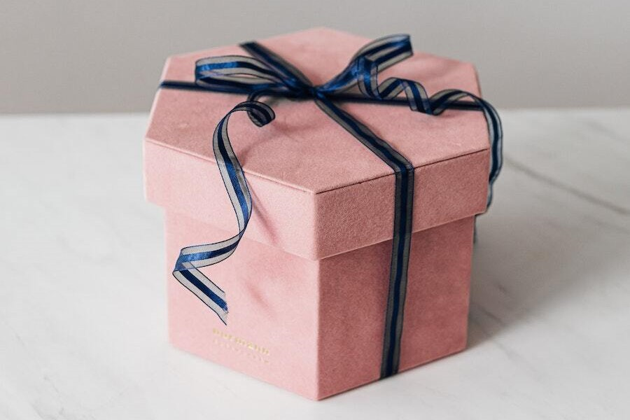 A pink, hexagonal gift box with a dark blue ribbon tied in a bow sits on a white surface.