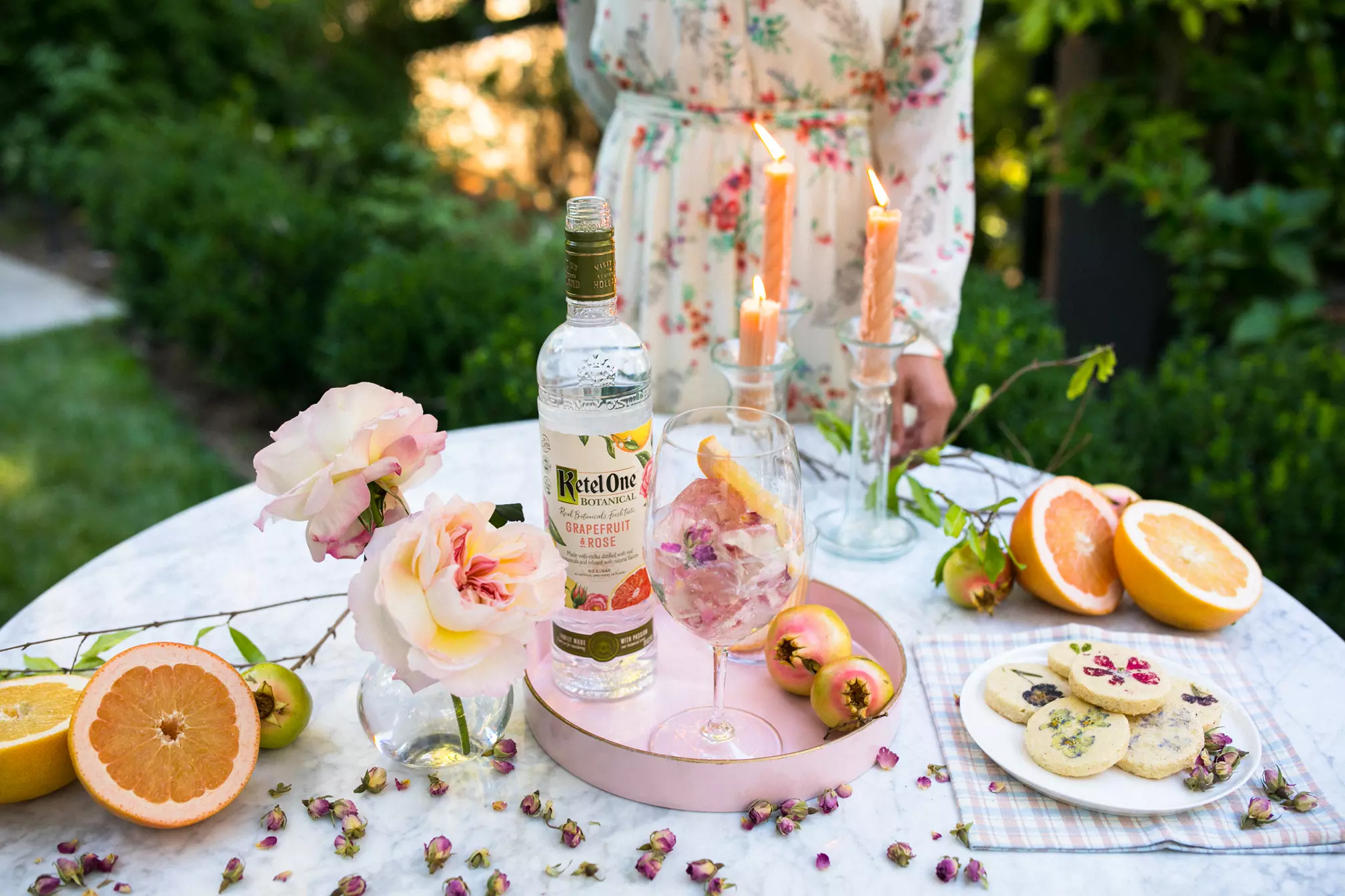 A round table outdoors holds a bottle of Ketel One Botanical Grapefruit & Rose, a cocktail, candles, grapefruit halves, cookies, and flowers. A person in a floral dress stands behind the table.