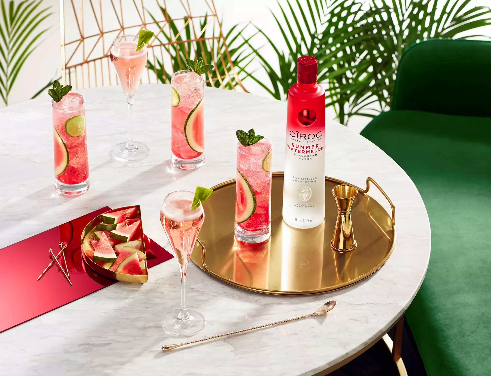 A marble table holds watermelon cocktails, a gold tray with a Ciroc Summer Watermelon bottle, sliced watermelon, cocktail tools, and glasses, with green plants in the background.