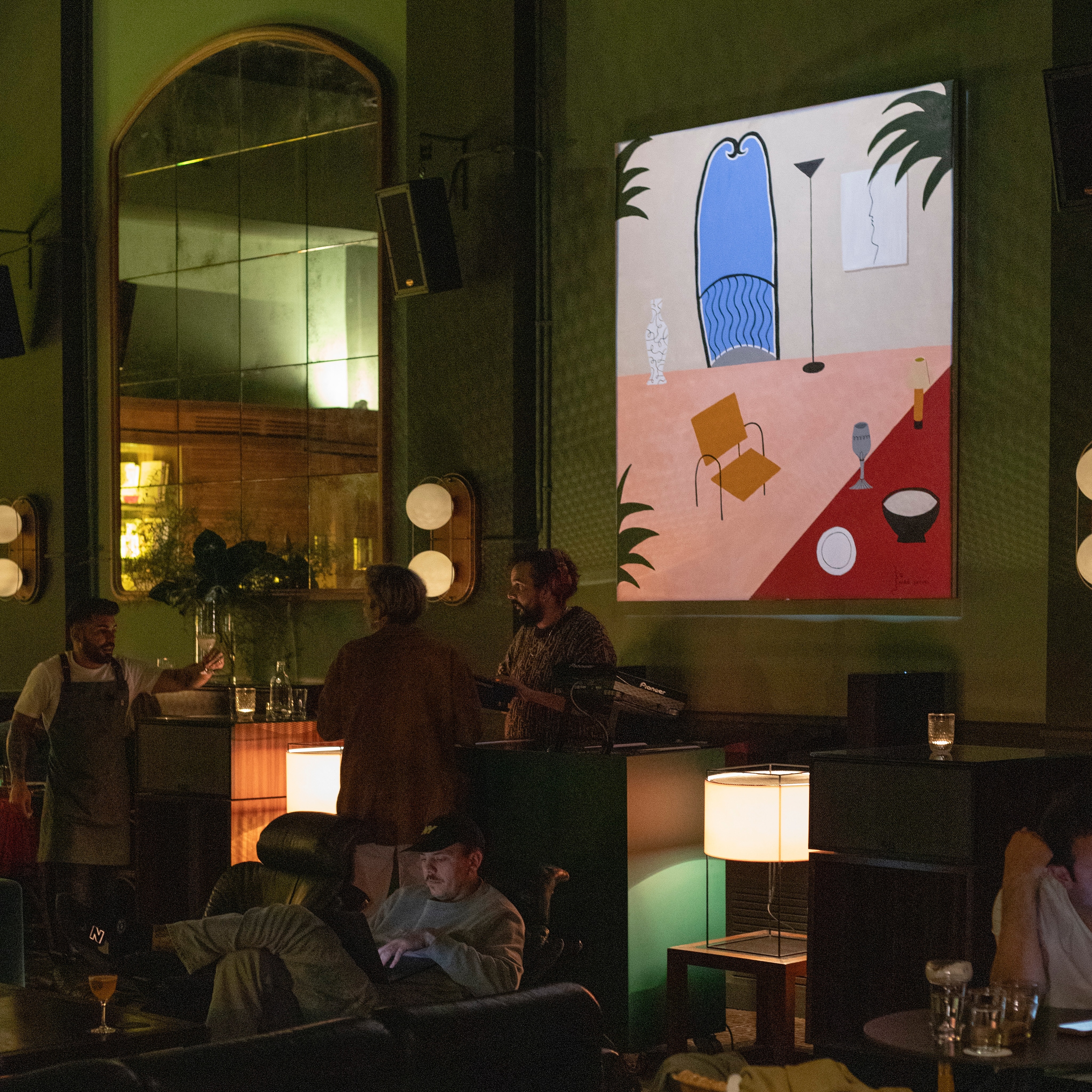 En El Chiringuito de Casa Bonay, un ambiente artístico y relajado destaca con iluminación cálida, espejos vintage y una obra de arte vibrante.