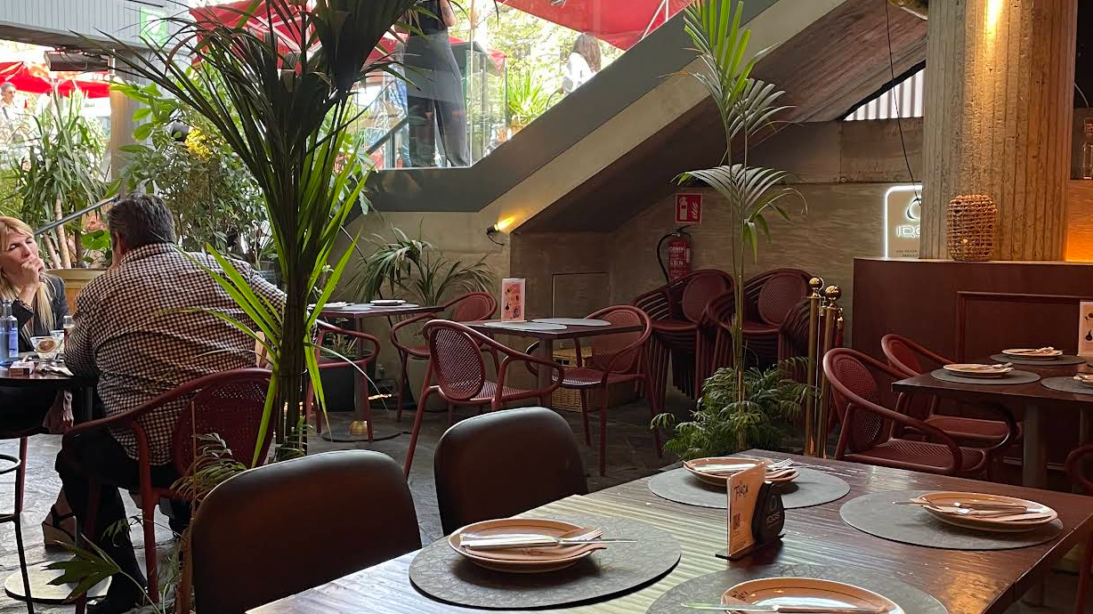 La imagen muestra un área de comedor exterior de La Flaca con un ambiente moderno y acogedor. Hay mesas de madera con platos y cubiertos, rodeadas de sillas rojas. Grandes plantas en macetas añaden un toque de verdor al espacio.