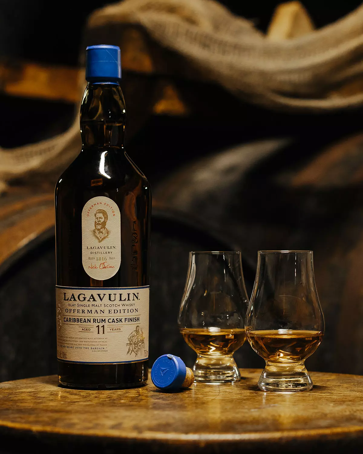 A bottle of Lagavulin 11 Year Old whisky, two filled whisky glasses, and a blue bottle cap are displayed on a wooden surface with barrels in the background.