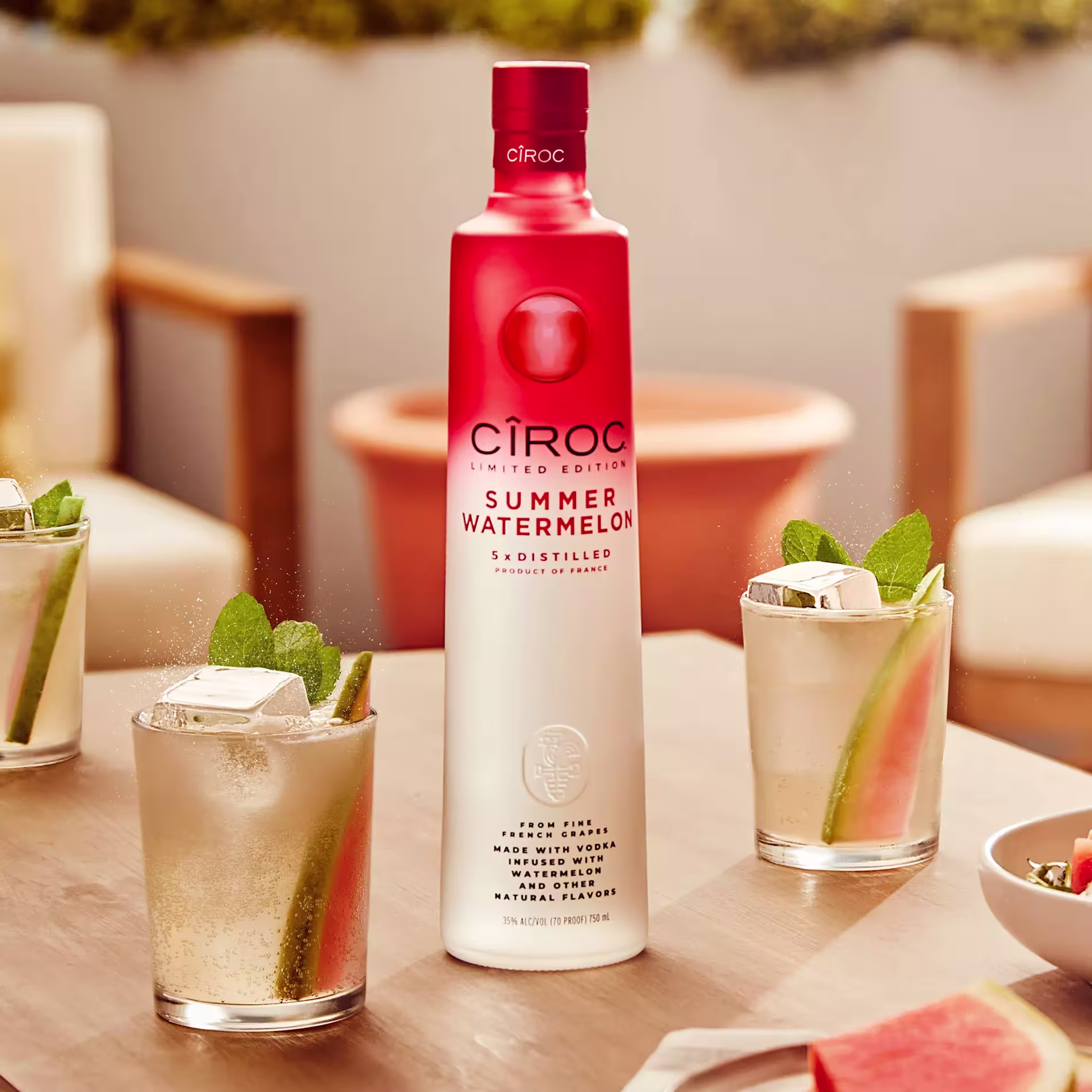 A bottle of Ciroc Summer Watermelon vodka stands on a table surrounded by three cocktails with ice, watermelon slices, and mint leaves.