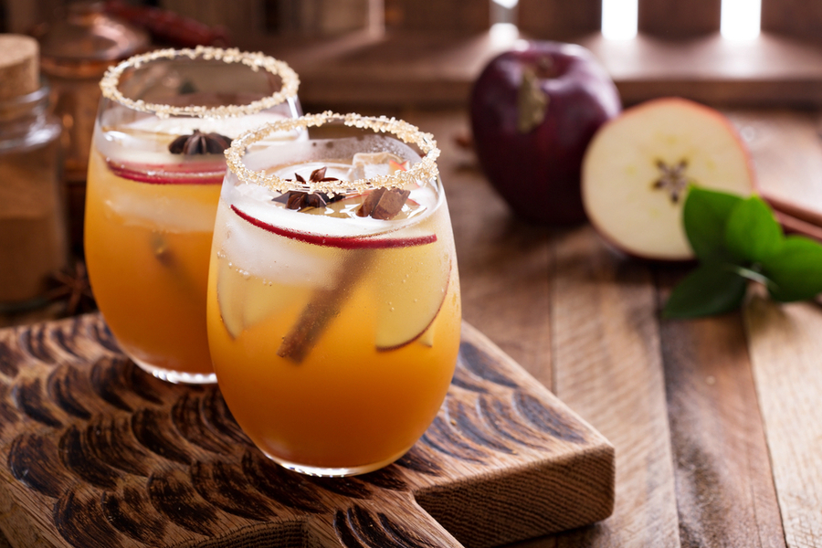 Two glasses of iced apple cider garnished with apple slices, star anise, and cinnamon sticks on a wooden board, with an apple and cinnamon stick in the background.