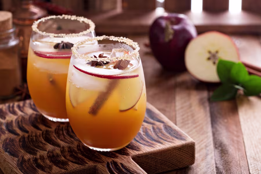 Two glasses of iced apple cider garnished with apple slices, star anise, and cinnamon sticks on a wooden board, with an apple and cinnamon stick in the background.