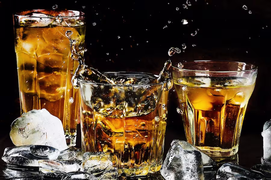 Three glasses of whiskey with ice cubes, with one glass showing a splash, surrounded by scattered ice cubes on a dark background.