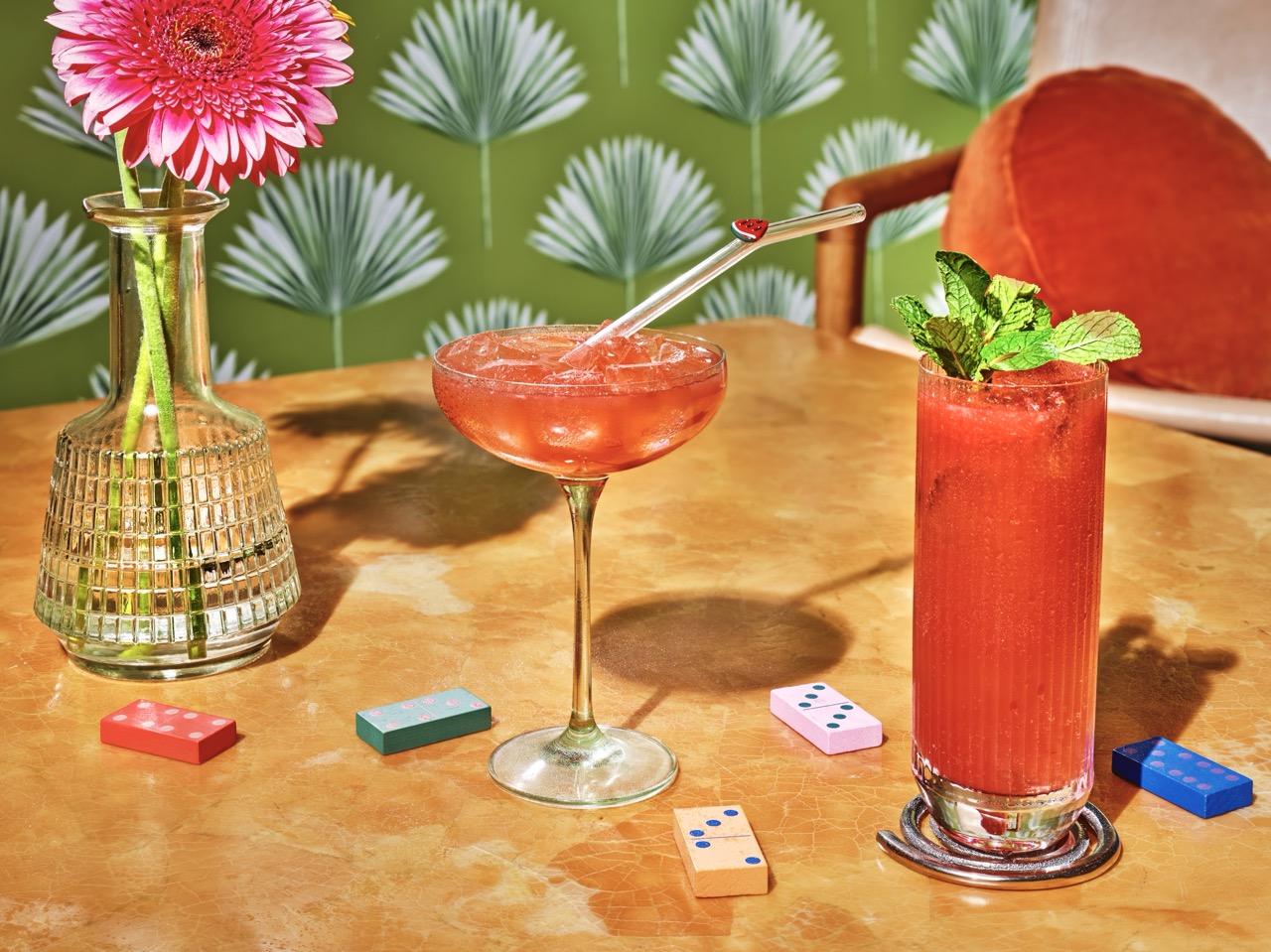 Two red cocktails sit on a marble table with scattered dominoes, a vase with a pink flower, and a green leaf-patterned wallpaper in the background.