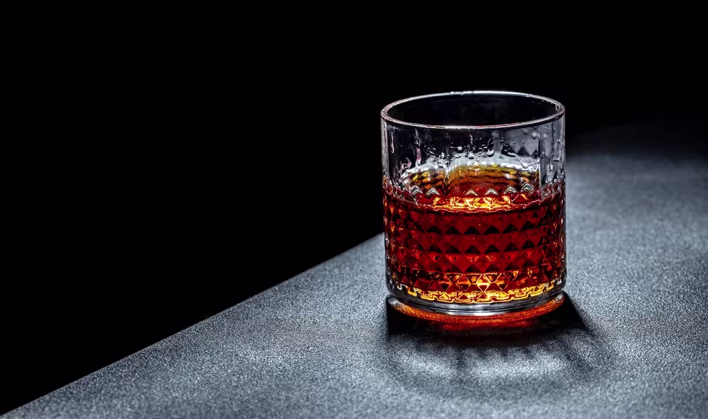A glass filled with a dark amber liquid, possibly whiskey or bourbon, sits on a textured dark surface against a black background.