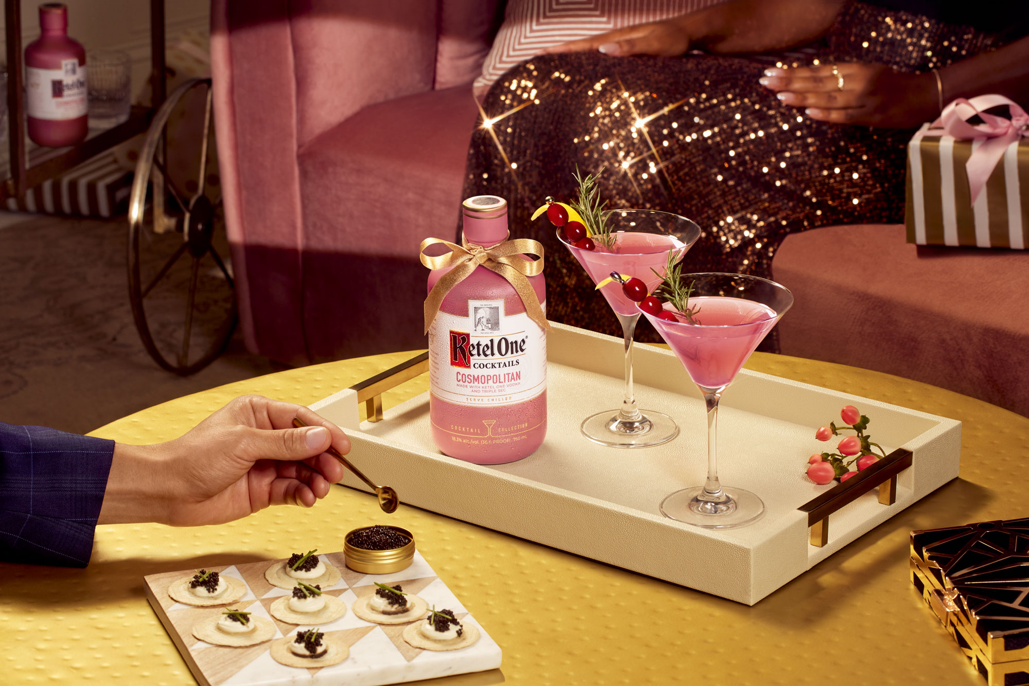 A tray with a bottle of Ketel One Cosmopolitan, two pink cocktails garnished with herbs and berries, and a plate of appetizers on a yellow table.