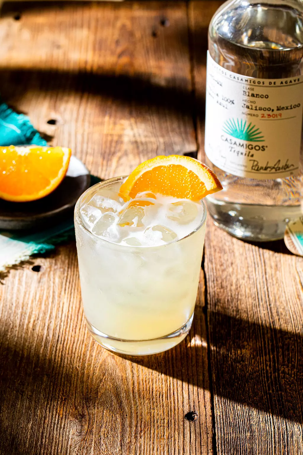 A glass of cocktail with ice and an orange wedge garnish sits on a wooden table next to a bottle of Casamigos tequila and an orange slice on a small plate.