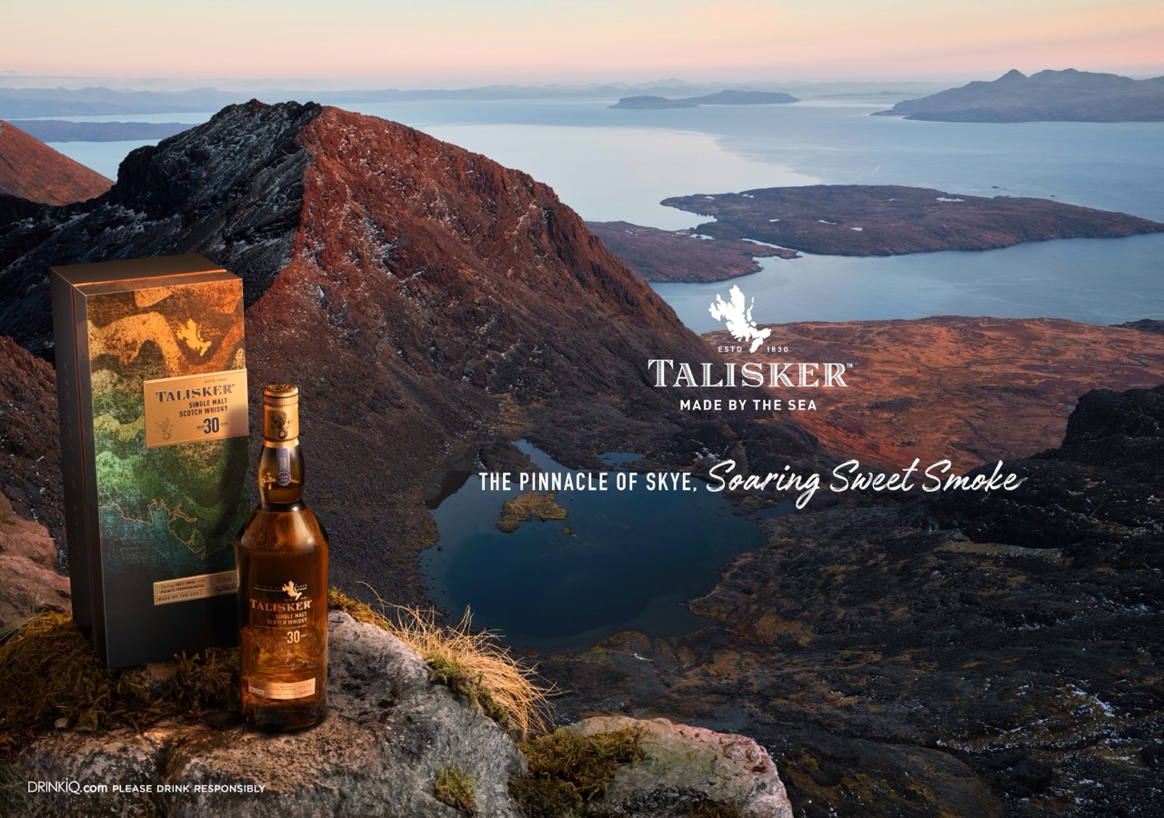 A bottle and box of Talisker 30-Year-Old Scotch whisky are placed on a grassy cliff with a scenic view of mountains, water, and sky in the background.