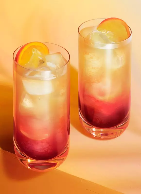 Two tall glasses of a layered red and yellow beverage with ice cubes and citrus slices, set against a warm orange background.