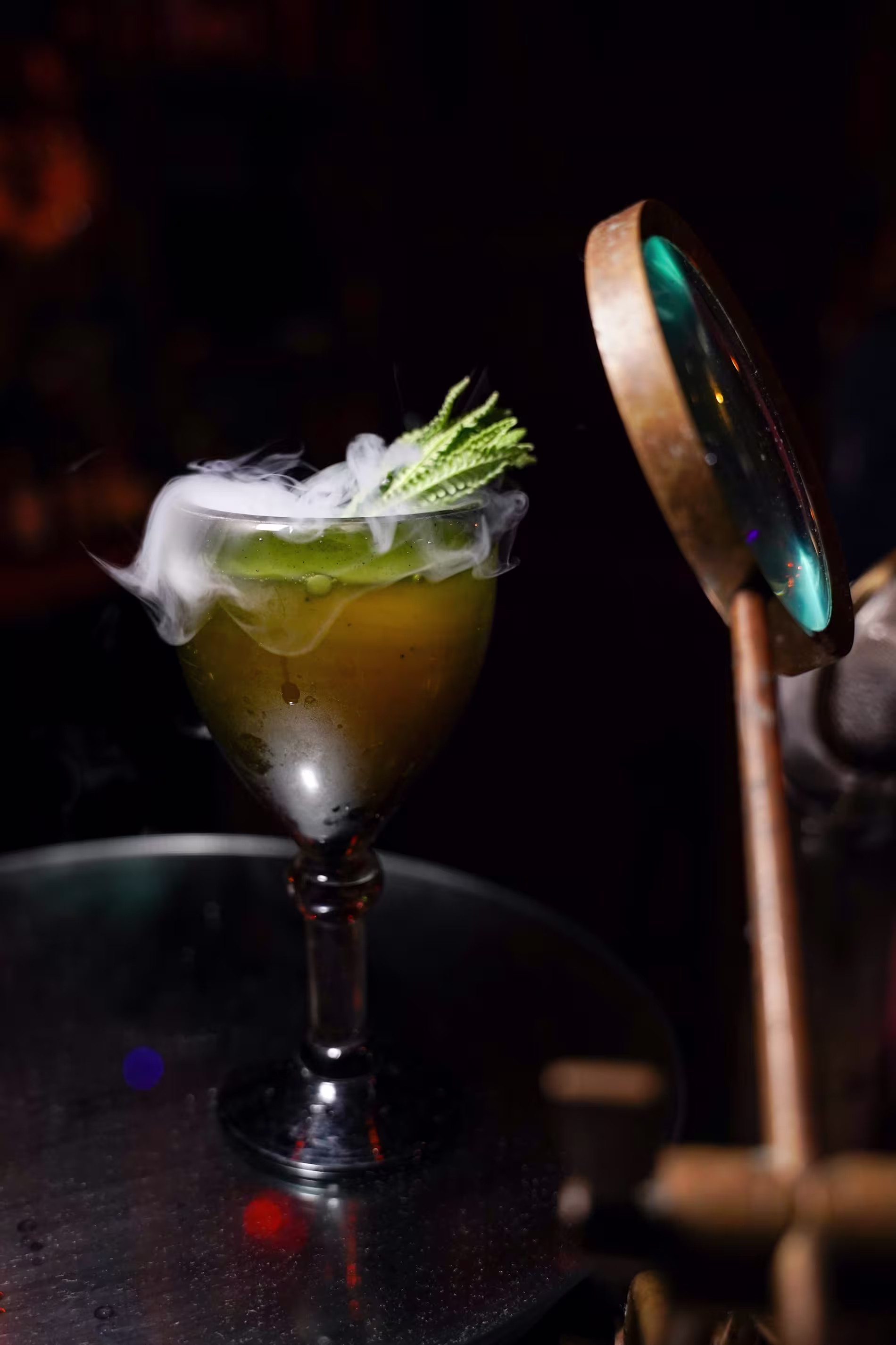A cocktail in a goblet emits wisps of smoke, garnished with herbs, and sits on a round table next to a magnifying glass.