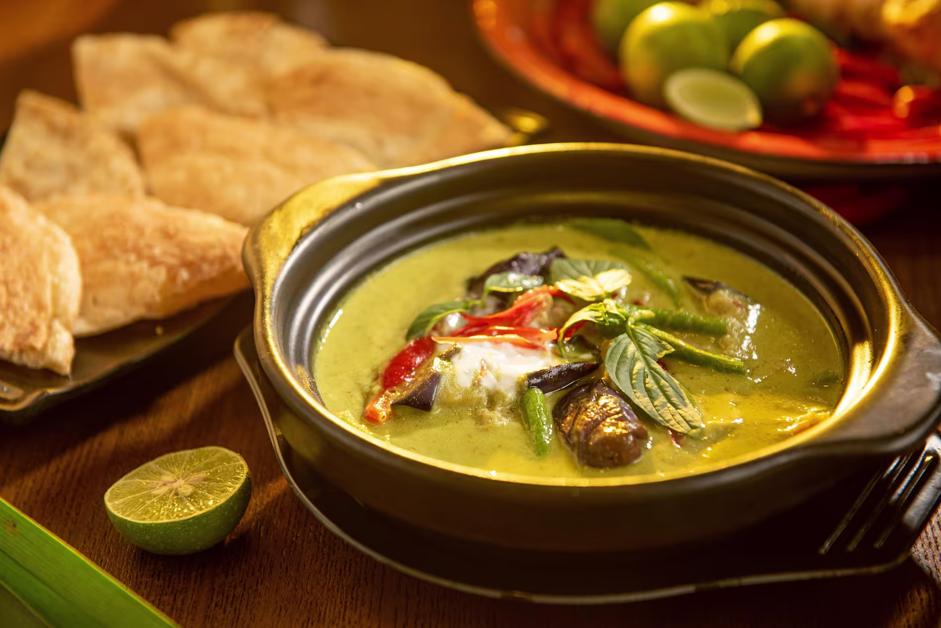 A bowl of green curry with vegetables and herbs, served with flatbread and lime on the side, placed on a wooden table.